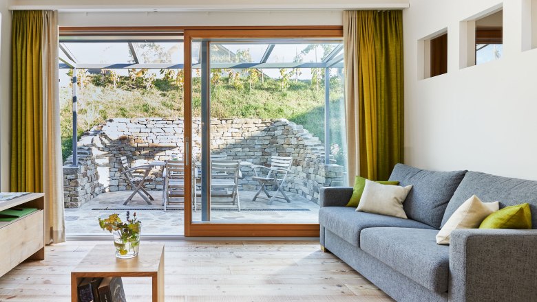 Weinviertler Kellerstöckl, © Andreas Hofer Pohled do interiéru moderního obývacího pokoje s šedou pohovkou, dřevěnou podlahou a velkými okny vedoucími na terasu s kamennou zdí a zahradním nábytkem.