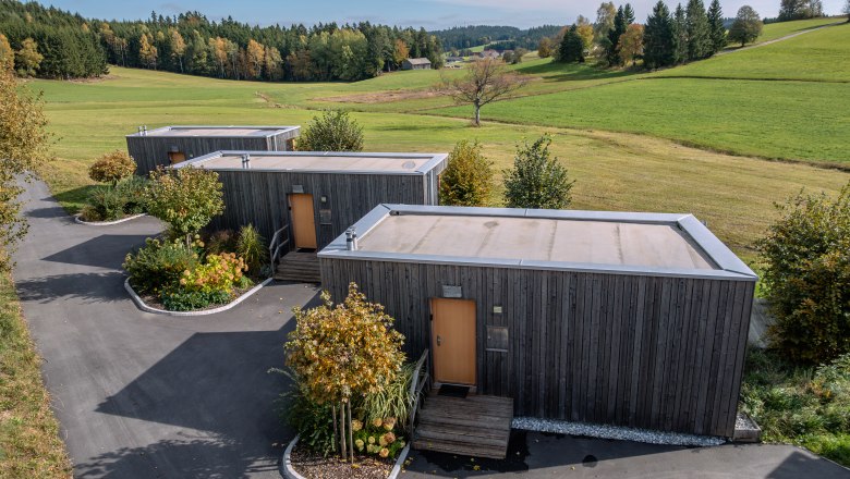 roo'n, © Niederösterreich Werbung / Maximilian Pawlikowsky Tři moderní dřevěné domy v zelené krajině se stromy a loukami.