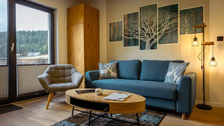 Ötscher Lodge, © Niederösterreich Werbung / Maximilian Pawlikowsky Útulný obývací pokoj s modrou pohovkou, křeslem, dřevěným stolem a výhledem na les z okna.