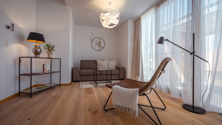 Apartmány Hausgnost, © Niederösterreich Werbung / Maximilian Pawlikowsky Moderně zařízený obývací pokoj s pohovkou, křeslem, stojací lampou a velkým oknem se závěsy.
