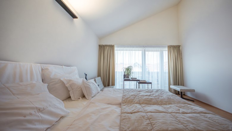 MÖRWALD, Relais Chateaux Hotel am Wagram, © Niederösterreich Werbung / Maximilian Pawlikowsky Moderní hotelový pokoj s velkou postelí, bílými polštáři a přehozem, světlými závěsy a oknem v pozadí.