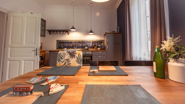 Dovolená v hotelu BAHNhof, © Niederösterreich Werbung / Maximilian Pawlikowsky Kuchyně s dřevěným stolem, hračka vlak, láhev a květiny na stole, moderní lampy a kuchyňské skříňky v pozadí.