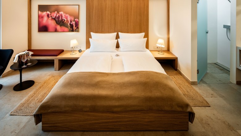 Penzion Flora, © Herbert Lehmann Moderně zařízený hotelový pokoj s manželskou postelí, nočními stolky, lampičkami a obrazem na stěně.