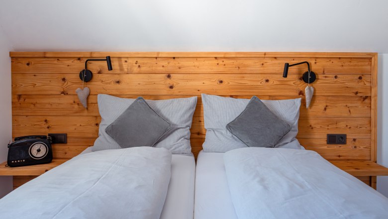 Leopold an der Ybbs, © © Niederösterreich Werbung/ Maximilian Pawlikowsky Útulná manželská postel s dřevěným opěradlem, šedými polštáři a přikrývkami. Vlevo retro rádio, vpravo lampa na čtení a dekorace srdce.