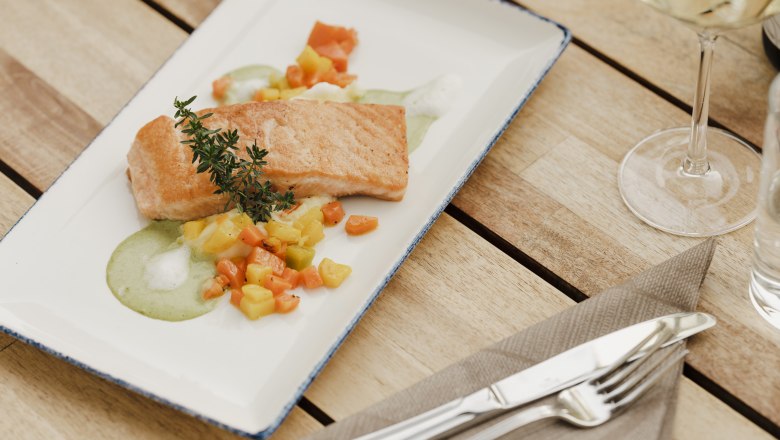 Losos na zelenině, © Niederösterreich Werbung/Michael Reidinger Talíř smaženého lososa se zeleninou a zelenou omáčkou, ozdobený snítkou bylinek, na dřevěném stole s příbory a skleničkou na víno.