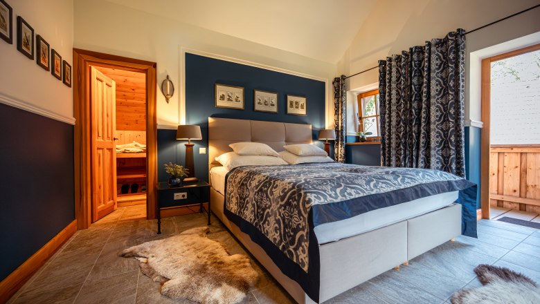 Lesnická chata Langschlägerwald, © Niederösterreich Werbung / Maximilian Pawlikowsky Útulná ložnice s manželskou postelí, modrými stěnami a dřevěnými prvky, která sousedí se saunou.