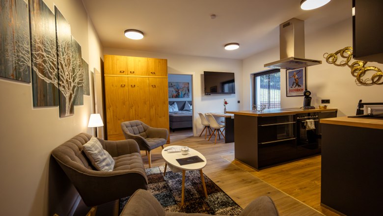 Ötscher Lodge, © Niederösterreich Werbung / Maximilian Pawlikowsky Moderní obývací pokoj s kuchyní, pohovkou, křesly a jídelním stolem. K vidění je světlý dřevěný nábytek a umělecká díla na stěnách.