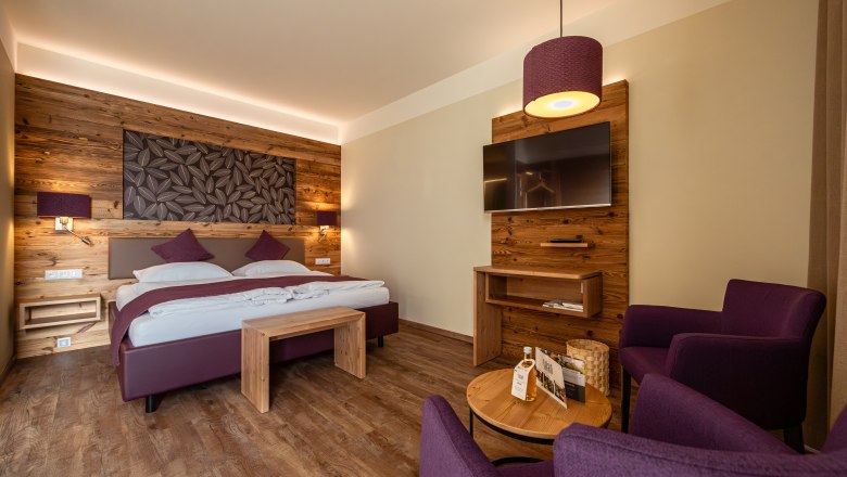 Postel ve Wies'n, © Niederösterreich Werbung / Maximilian Pawlikowsky Útulný hotelový pokoj s dřevěnými stěnami, manželskou postelí, TV a fialovými křesly.