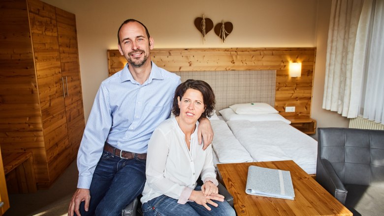 Pokoje na farmě Dissauer family Rosinger, © Familie Rosinger Pár sedí v útulném pokoji s dřevěným nábytkem a manželskou postelí.