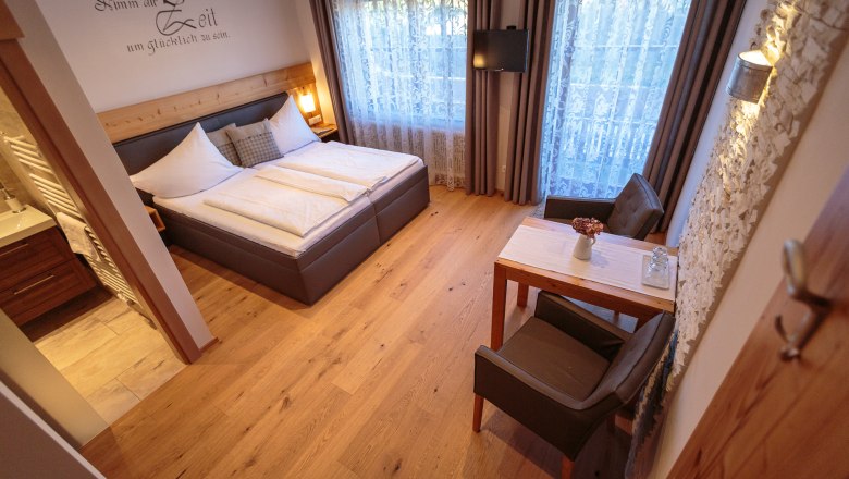 Dvoulůžkový pokoj, © rosinger Útulný dvoulůžkový pokoj s dřevěnou podlahou, manželskou postelí, malým stolkem a židlemi, ozdobnými závěsy a TV na stěně.