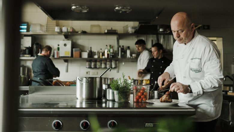 Manfred Hausgnost v kuchyni, © Niederösterreich Werbung/Michael Reidinger Kuchaři pracují v profesionální kuchyni, jeden krájí maso.