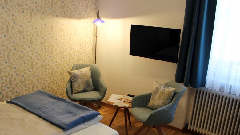 Room with bed, table with two chairs, TV, window with blue curtains and floor lamp., © Magdalena Donhauser Room with bed, table with two chairs, TV, window with blue curtains and floor lamp., © Magdalena Donhauser