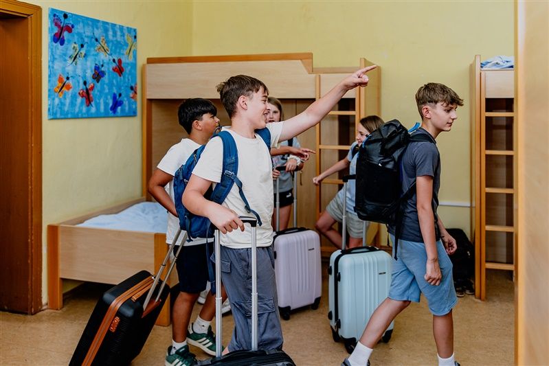 Skupina dětí s kufry v noclehárně s palandami.