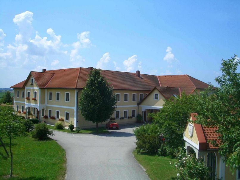 Velký statek se žlutými zdmi a červenými střechami, obklopený stromy a silnicí.