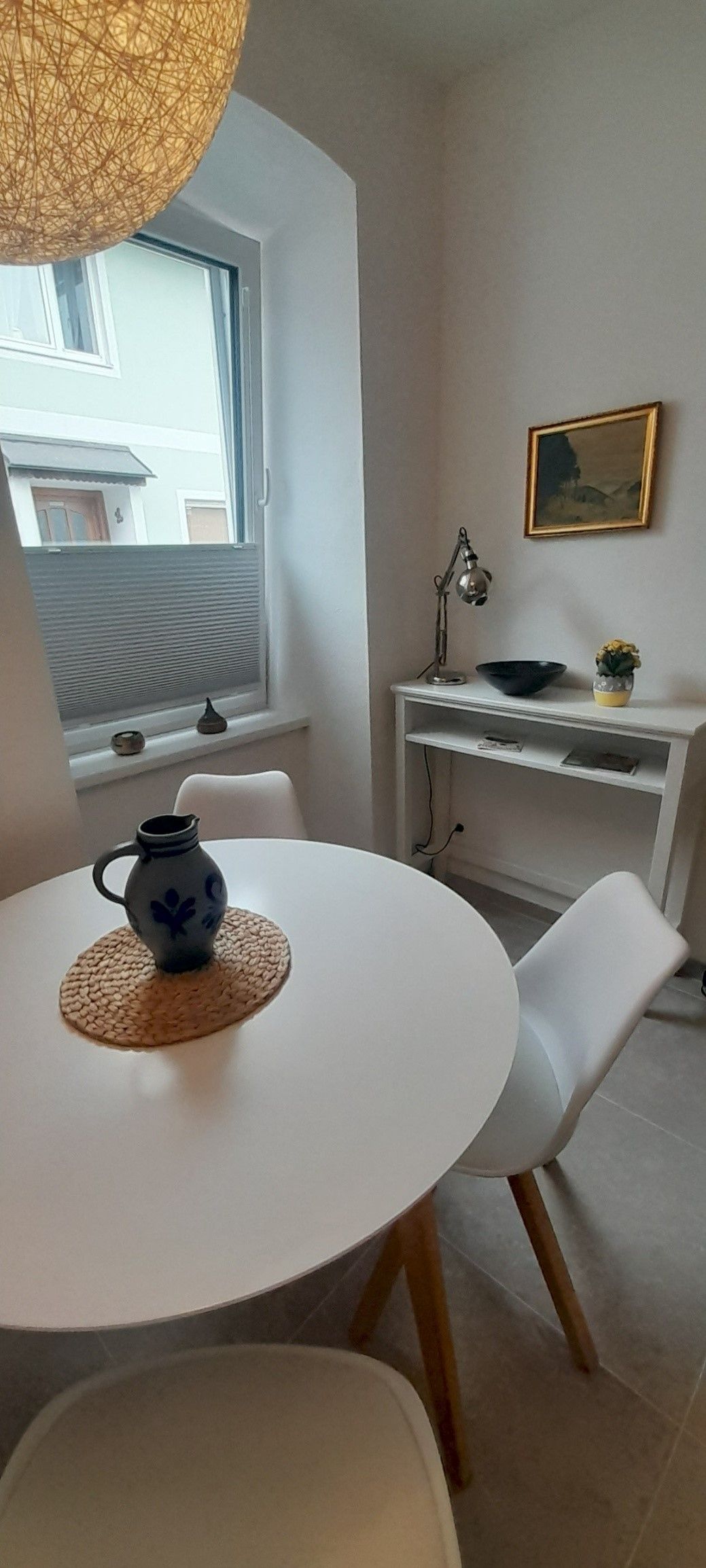 Malá jídelna s kulatým bílým stolem, dvěma bílými židlemi a ozdobnou vázou. V pozadí okno a malý stolek s lampou.
