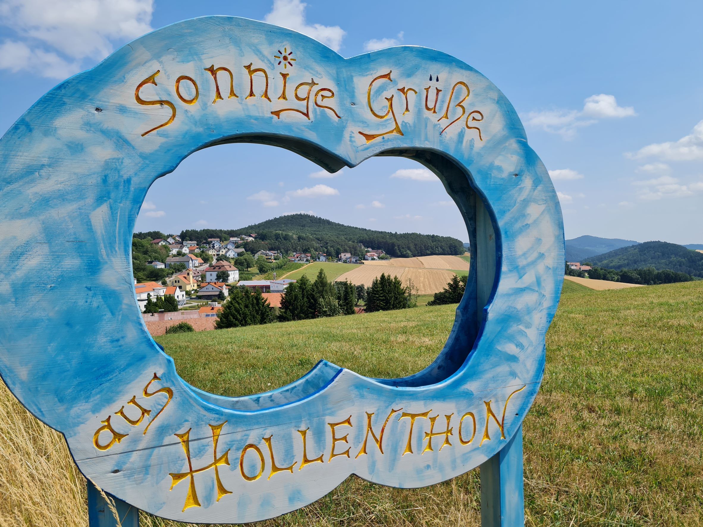 Modrý dřevěný rám ve tvaru mraku s nápisem "Sunny greetings from Hollenthon" na pozadí krajiny.