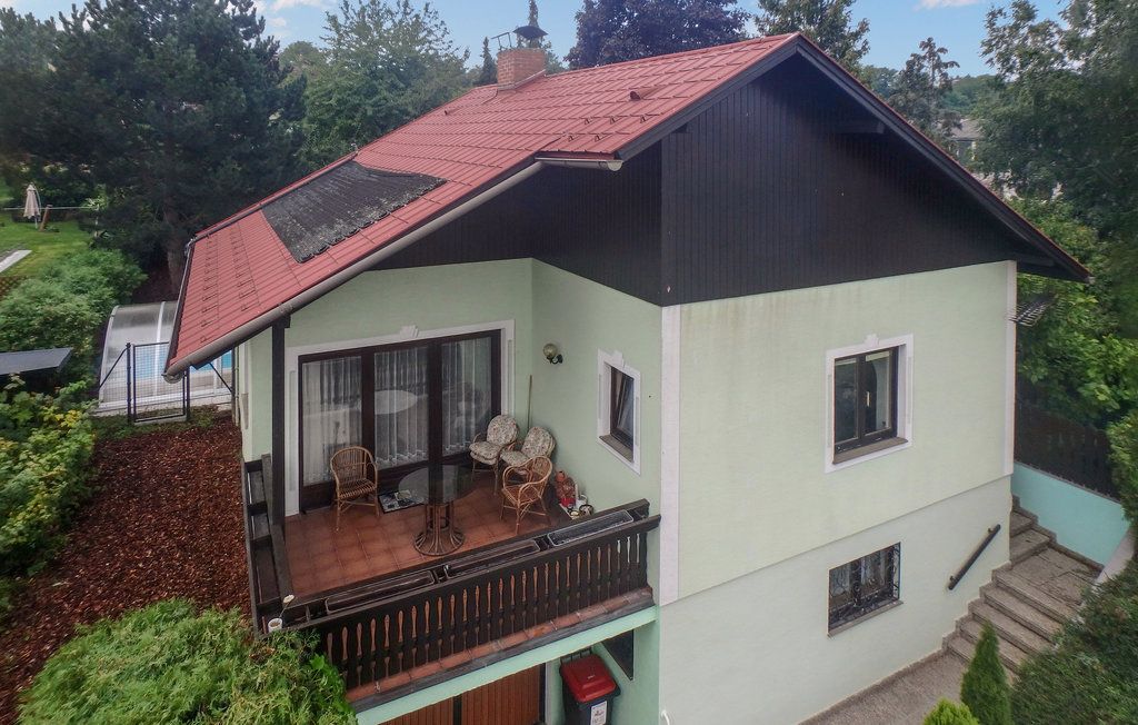 Dvoupodlažní dům s červenou střechou a balkonem, obklopený stromy.