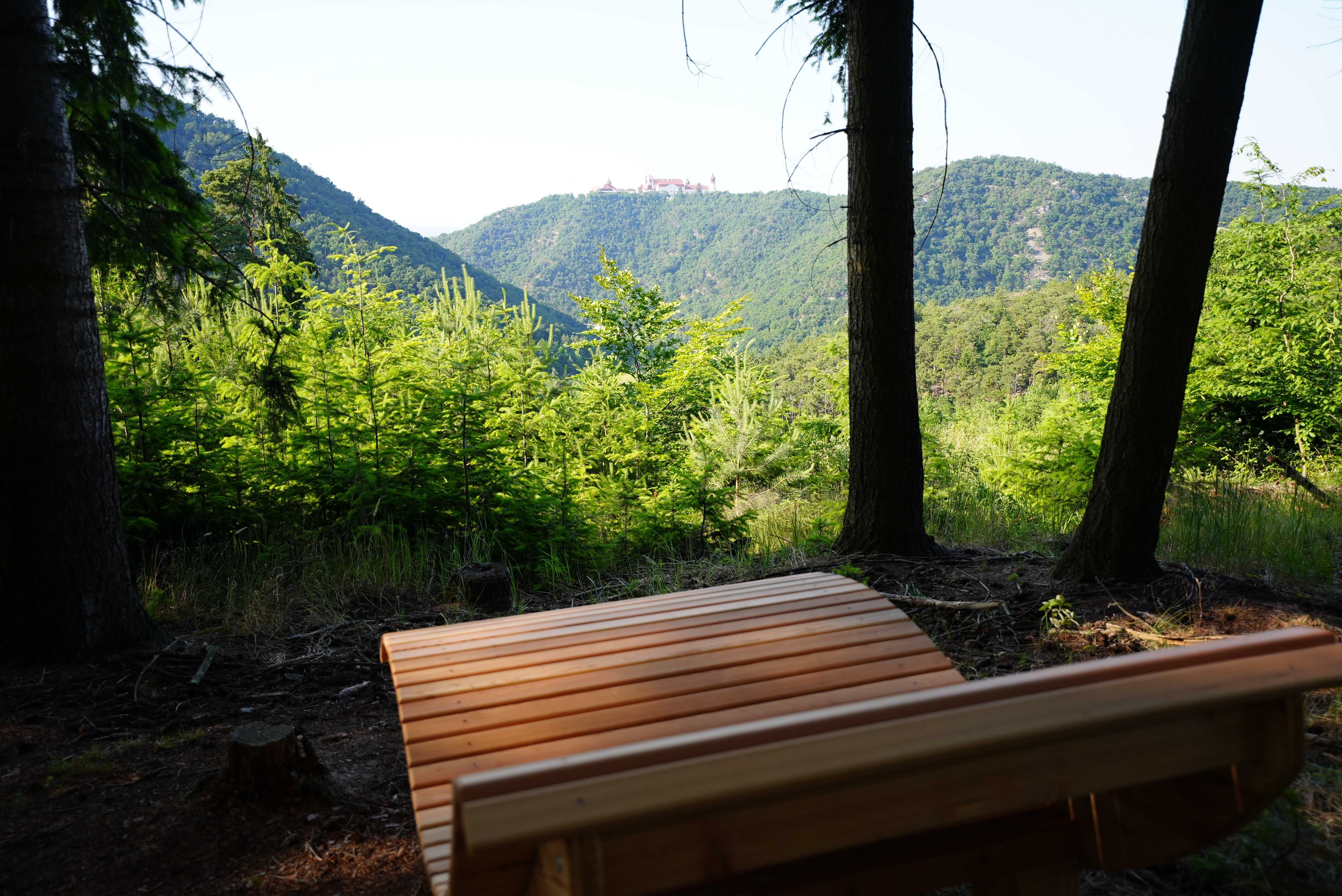 Dřevěná lavička v popředí s výhledem na zalesněné kopce a klášter v dálce.