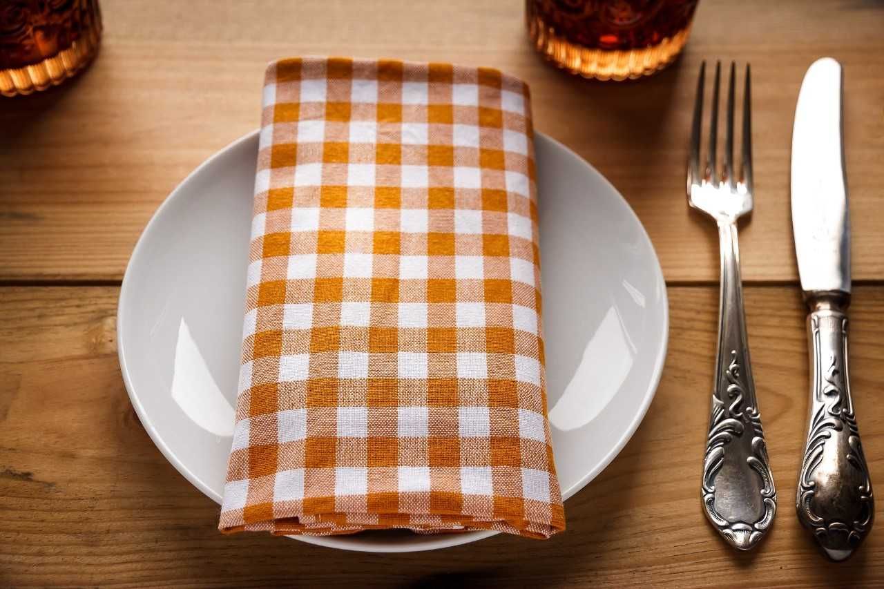 Talíř s oranžovo-bílým kostkovaným ubrouskem, vedle něj na dřevěném stole příbor.