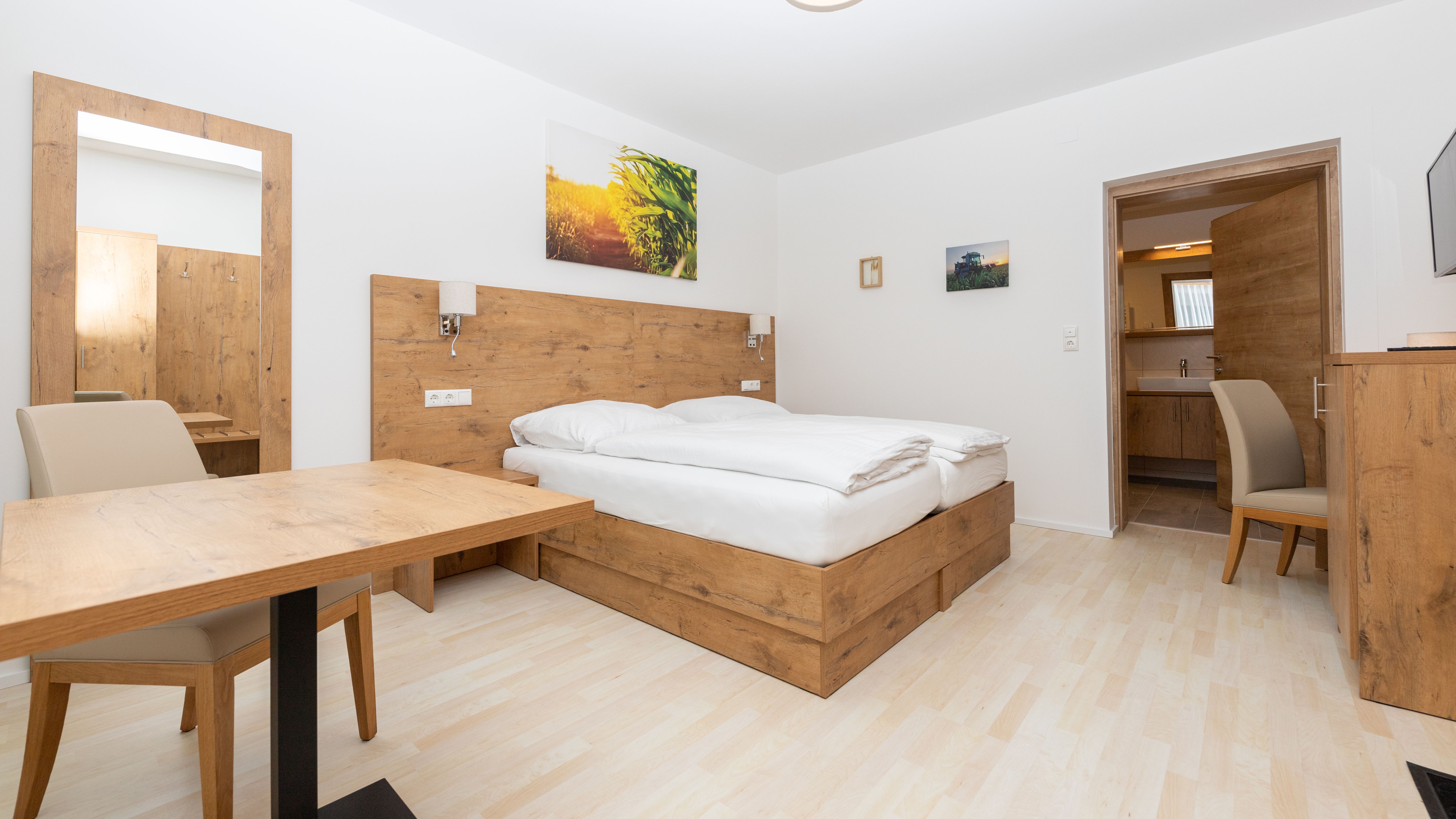 Moderní ložnice s dřevěným nábytkem, manželskou postelí, stolem a židlí. Zrcadlo a obrazy na stěně, dveře vedoucí do koupelny.