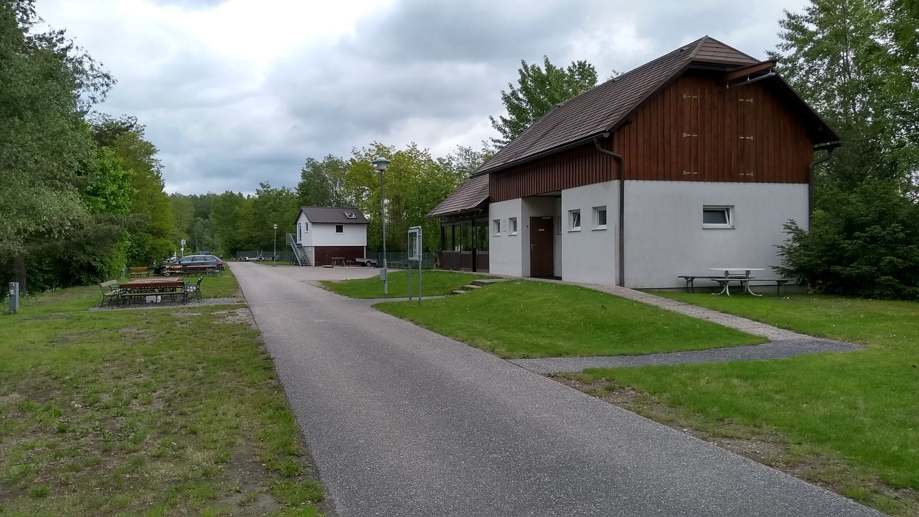Cesta vede kolem bílé budovy s dřevěným obložením, obklopené stromy a loukami, pod zamračenou oblohou.