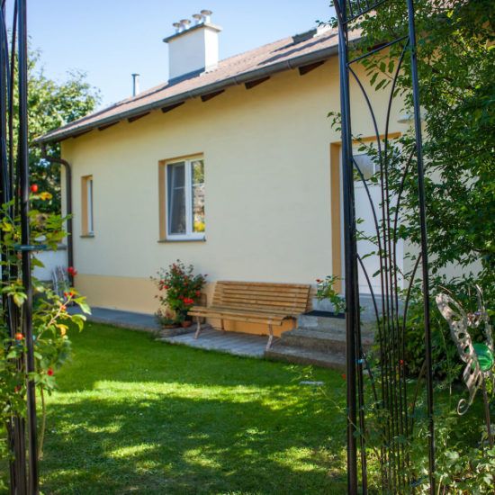Žlutý domek se zahradní lavičkou a květinami v popředí.