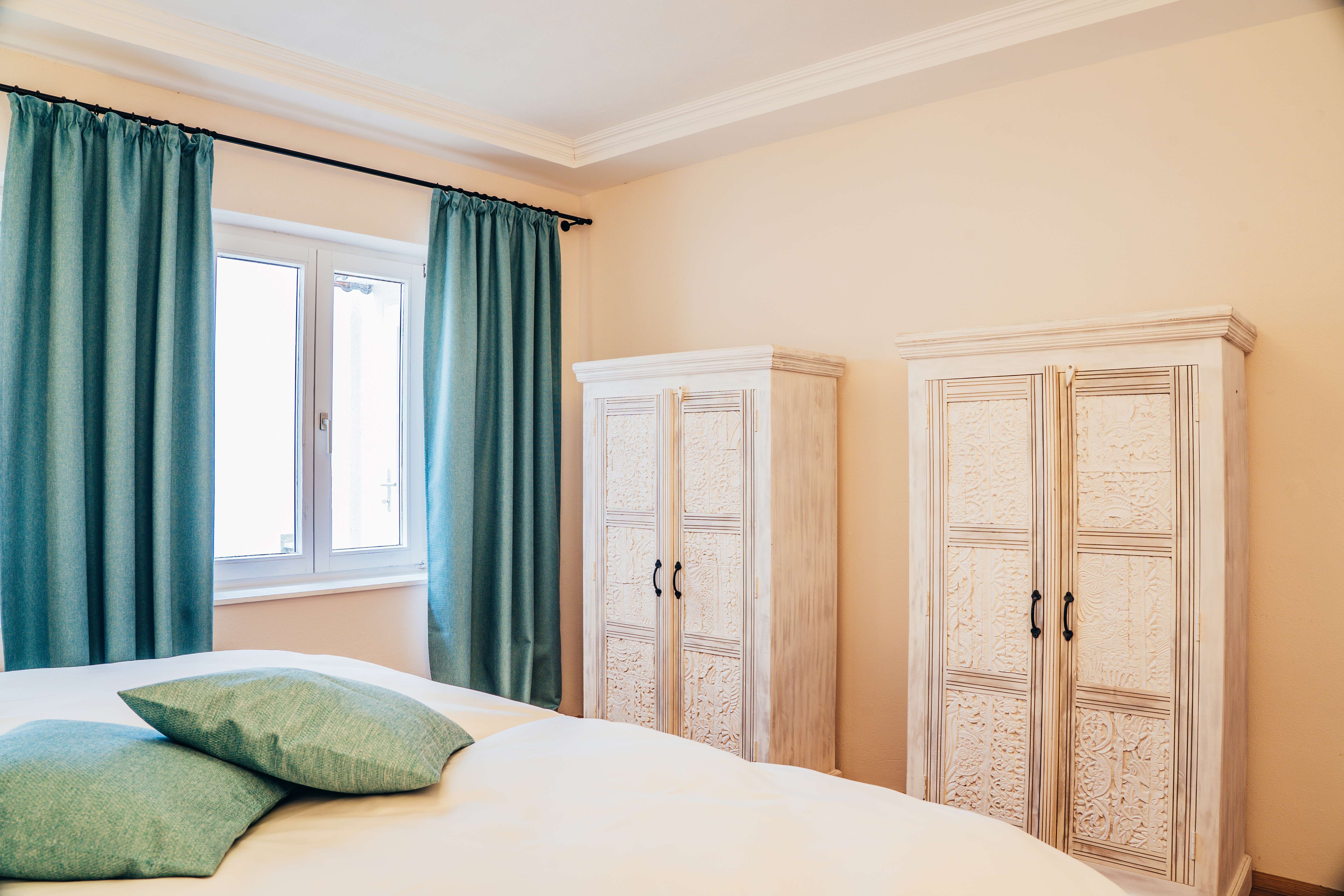 Ložnice se dvěma bílými skříněmi, tyrkysovými závěsy a polštáři.