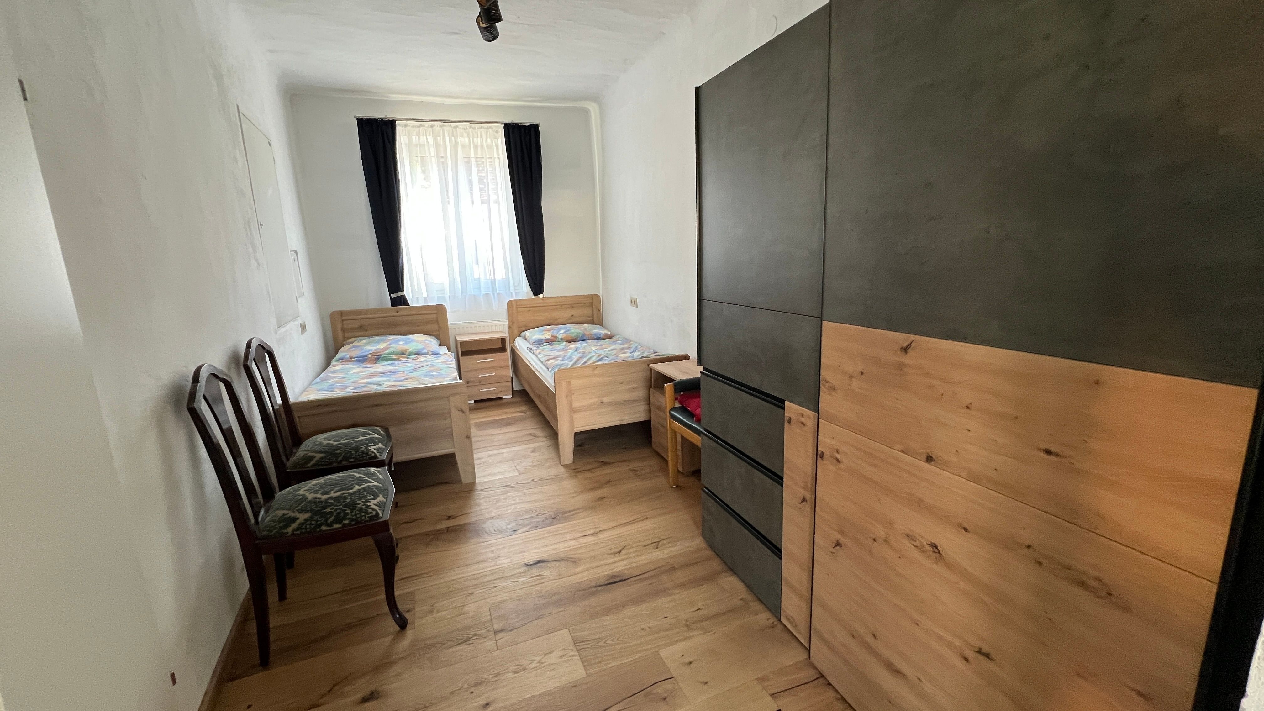 Malá ložnice se dvěma samostatnými lůžky, šatní skříní a dvěma židlemi. Okno se závěsy propouští světlo.
