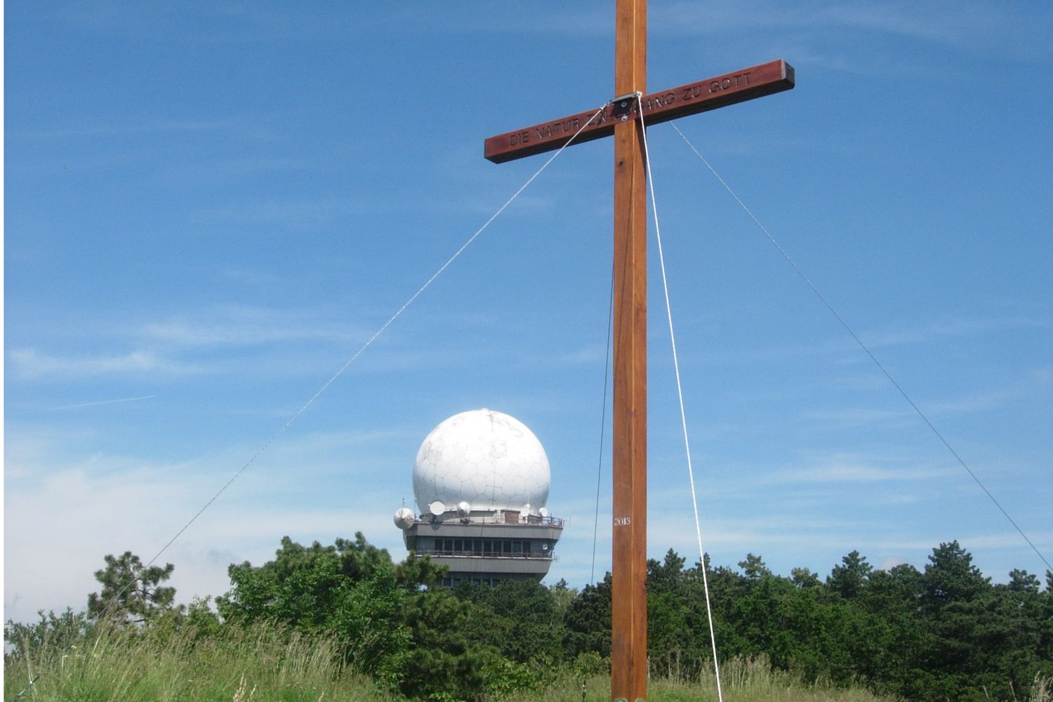 Dřevěný kříž před radarovou kopulí na Buschbergu, obklopený stromy a modrou oblohou.