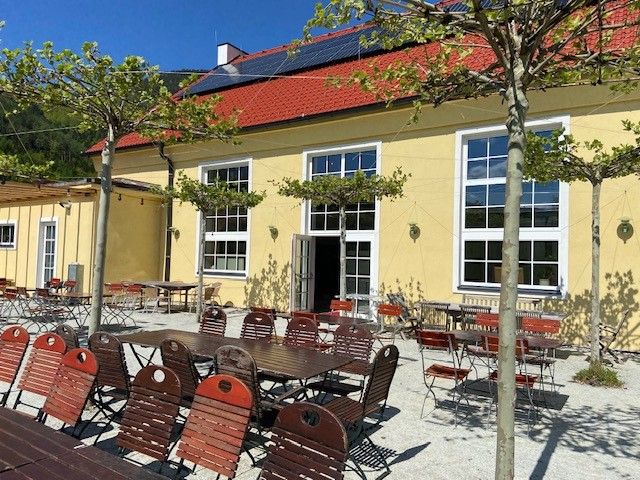 Venkovní prostor restaurace se žlutou budovou, červenými židlemi a stoly pod stromy.