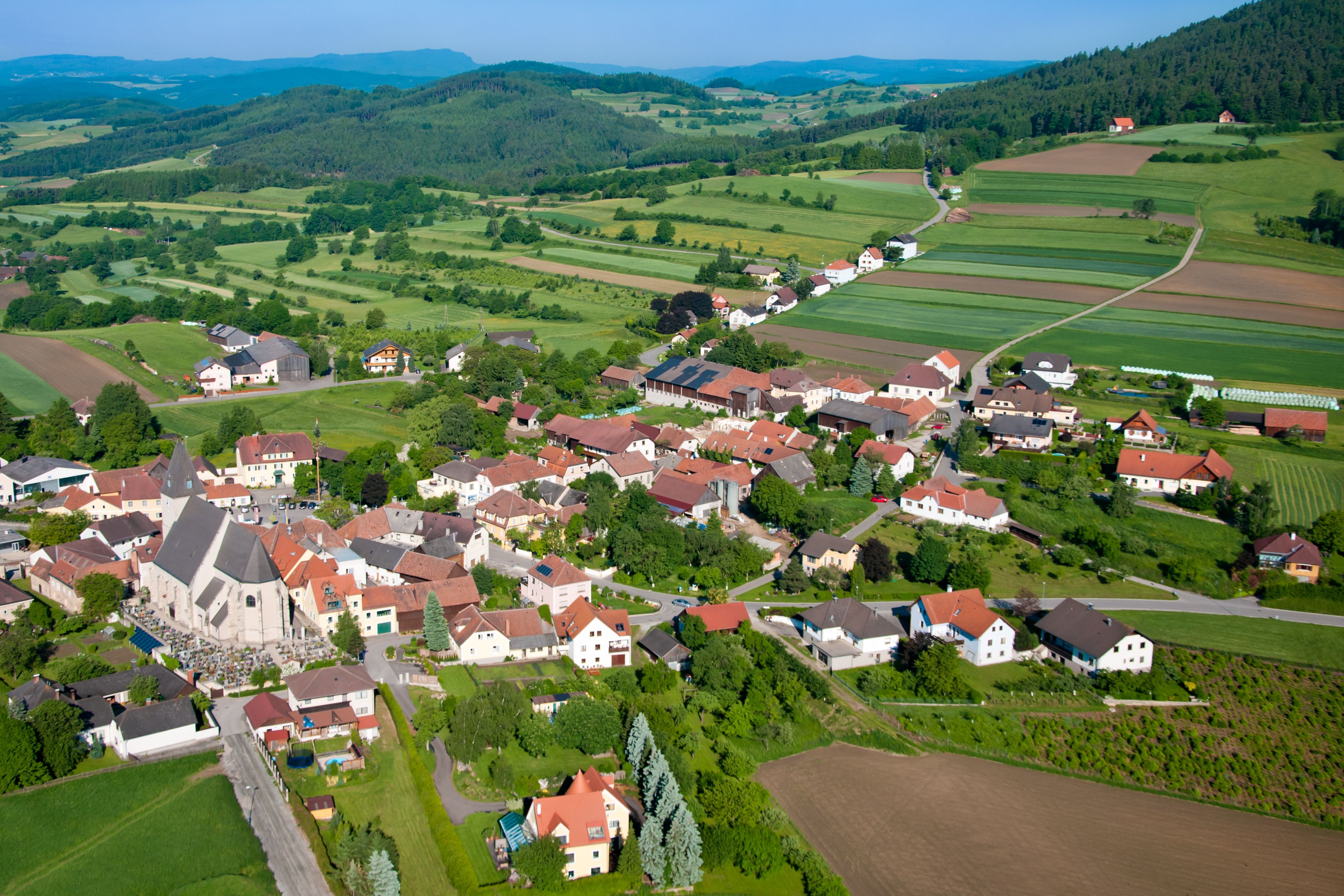 Letecký pohled na vesnici s kostelem, obklopenou zelenými poli a kopci.