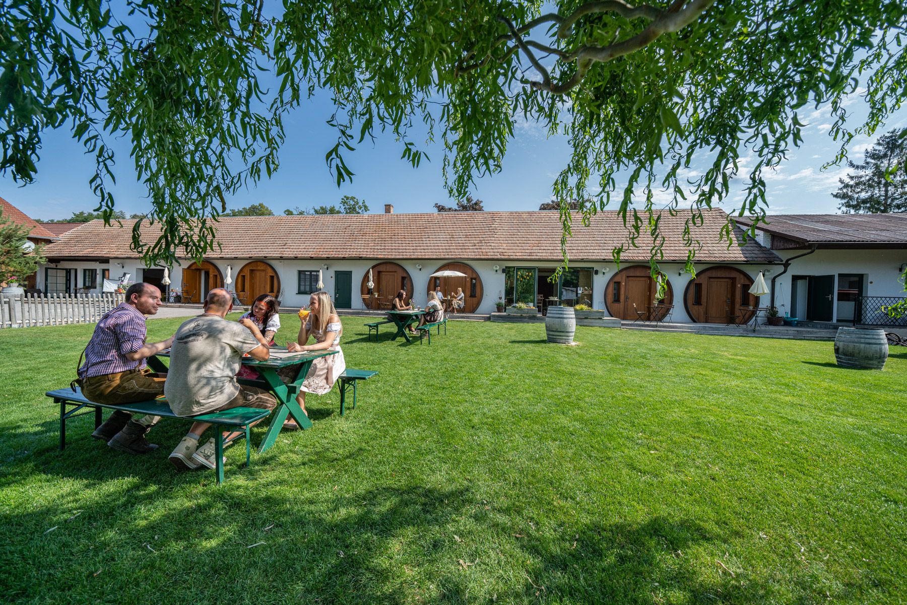 Lidé sedí u stolu na trávníku před budovou s kulatými dveřmi.