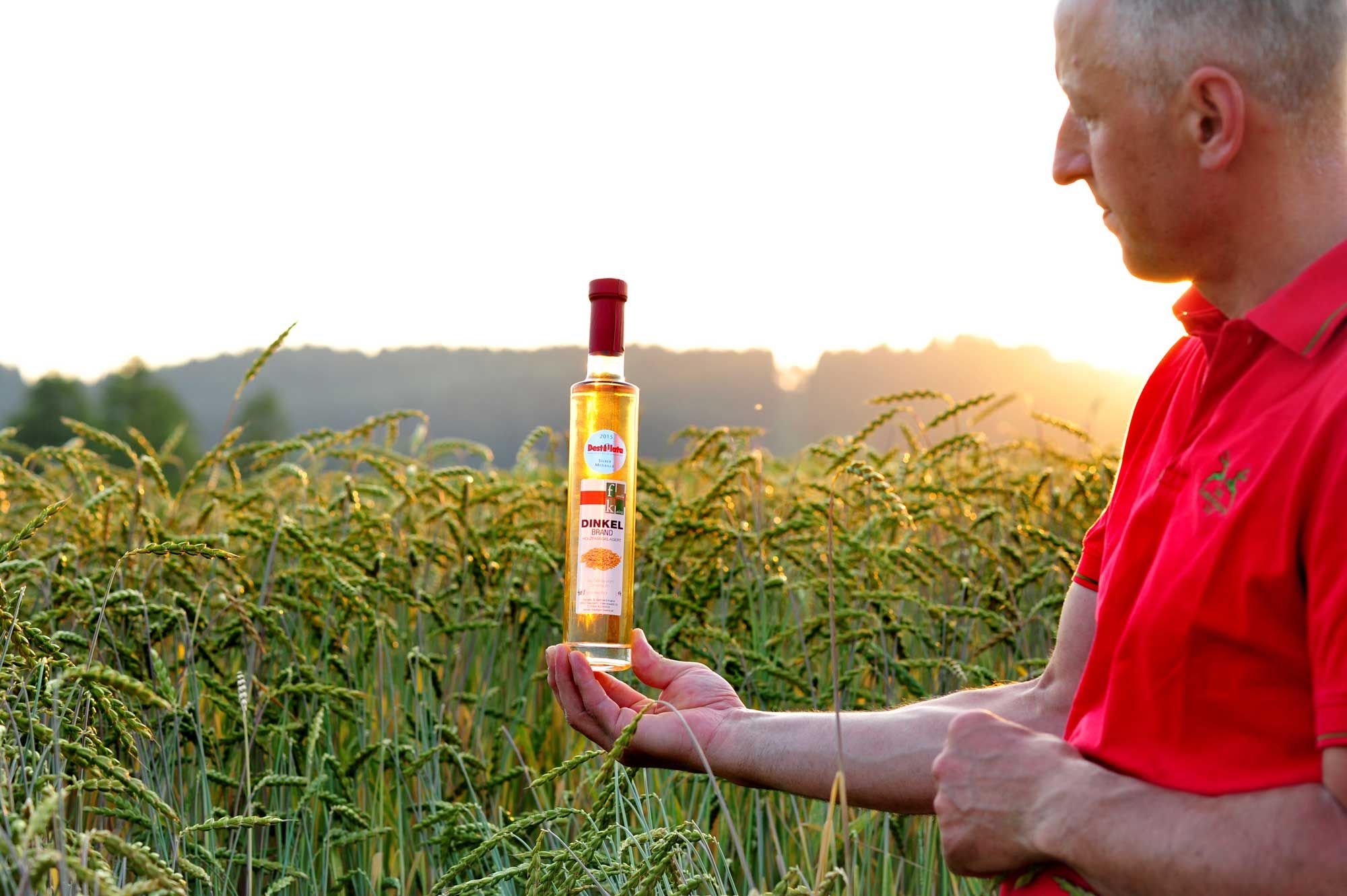 Muž v červené košili drží láhev špaldové pálenky v kukuřičném poli při západu slunce.