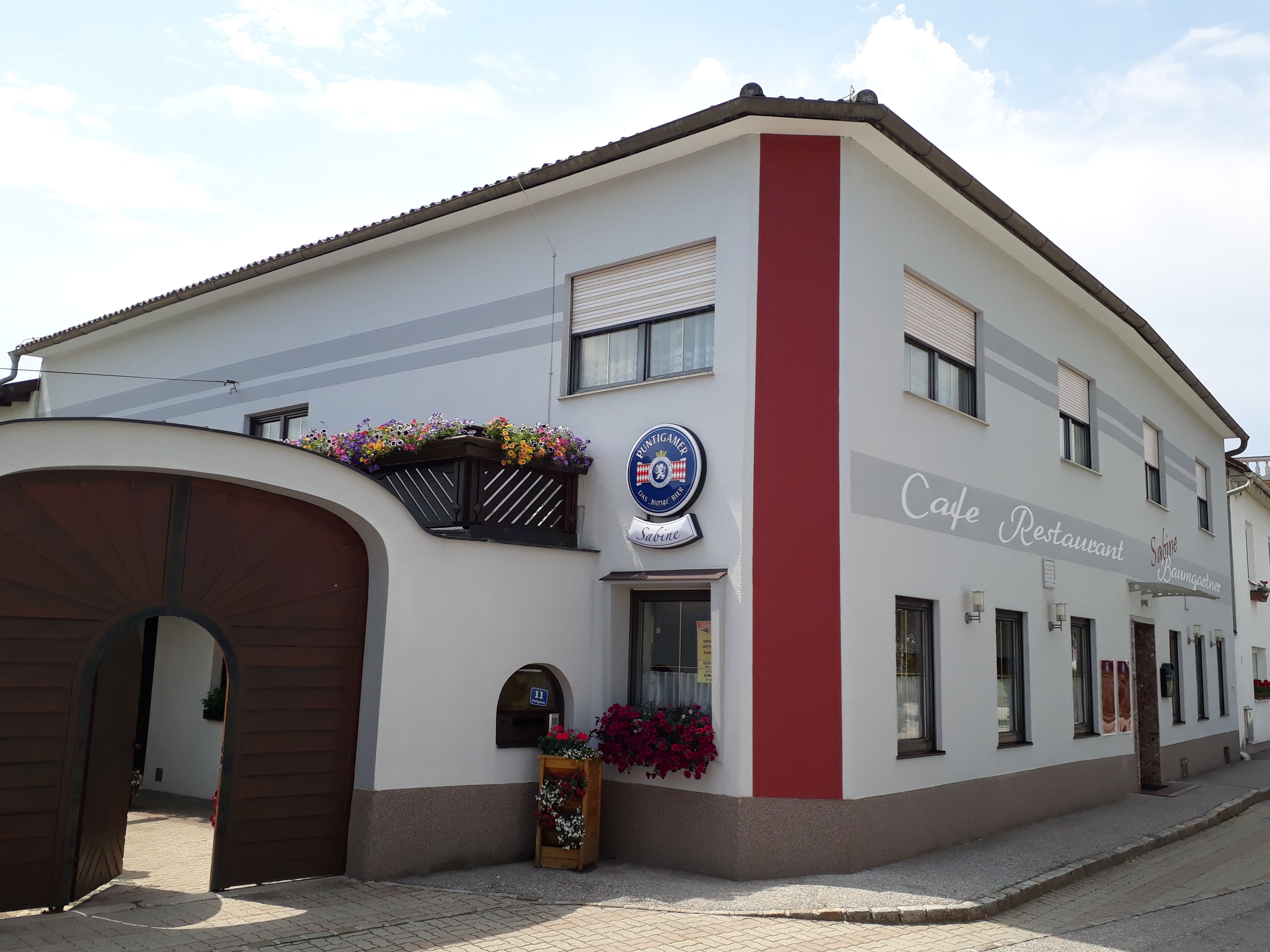 Pohled zvenčí na kavárnu Café Sabine s dřevěnou bránou, bílou a červenou fasádou a nápisem "Café-Restaurant".