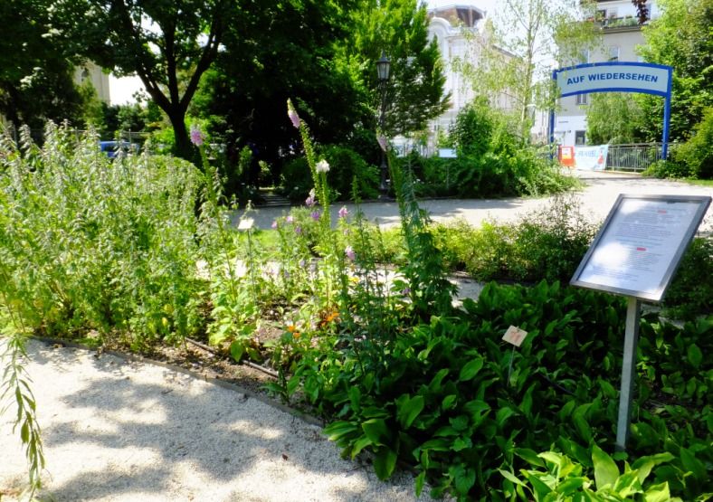 Upravená zahrada s rostlinami a cedulí, v pozadí brána s nápisem "Auf Wiedersehen".