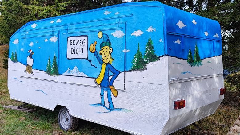 Barevně vymalovaný karavan se zimní krajinou a postavou s nápisem "Hejbejte se!".