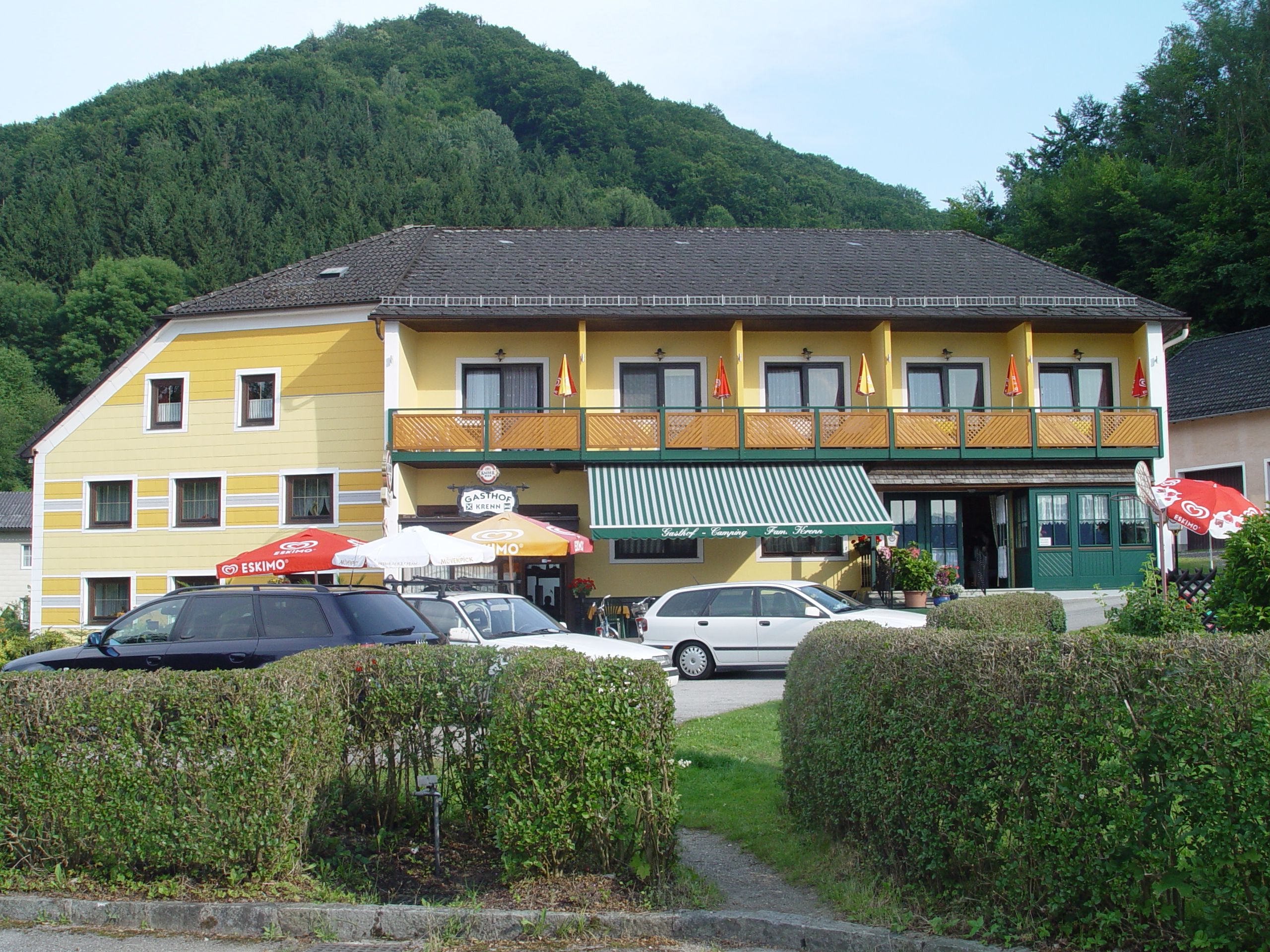 Žlutý penzion s balkonem a slunečníky před zalesněným kopcem.
