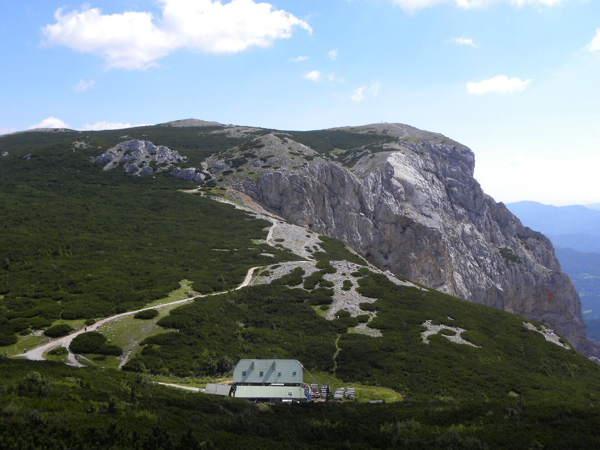 Horská krajina na Raxu s pozoruhodnými skalními útvary, turistickými stezkami a horskou chatou pod modrou oblohou.