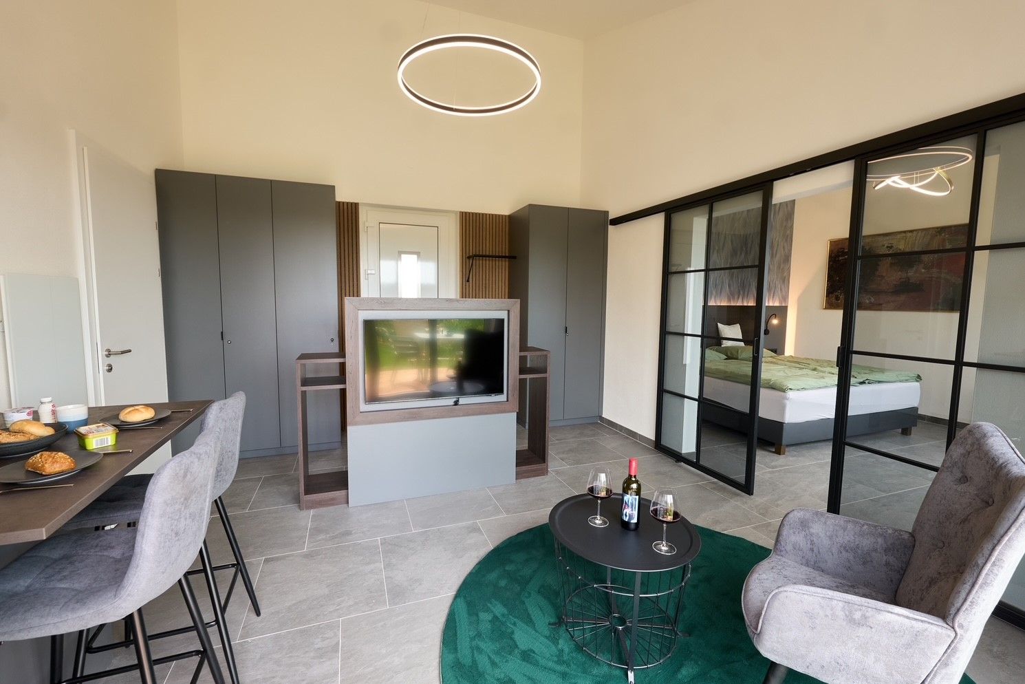 Moderní byt s jídelním stolem, TV, šedými skříněmi a posuvnými skleněnými dveřmi do ložnice.