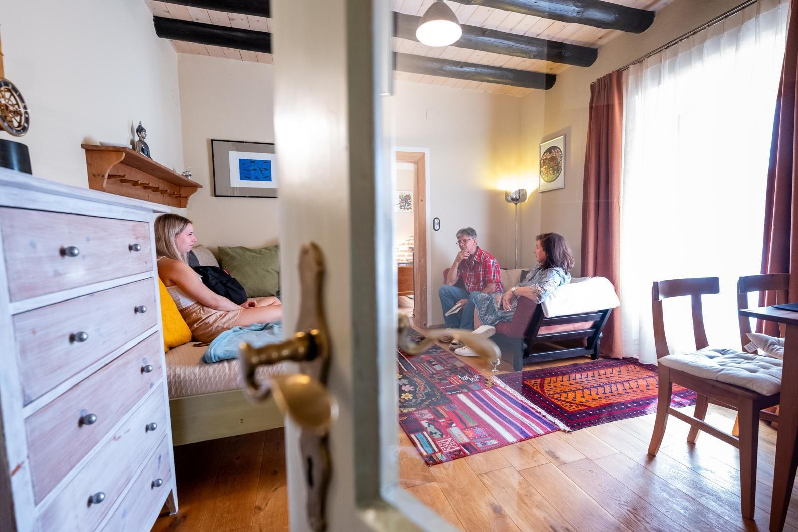 V útulném obývacím pokoji s dřevěnou podlahou a barevným kobercem sedí tři osoby.