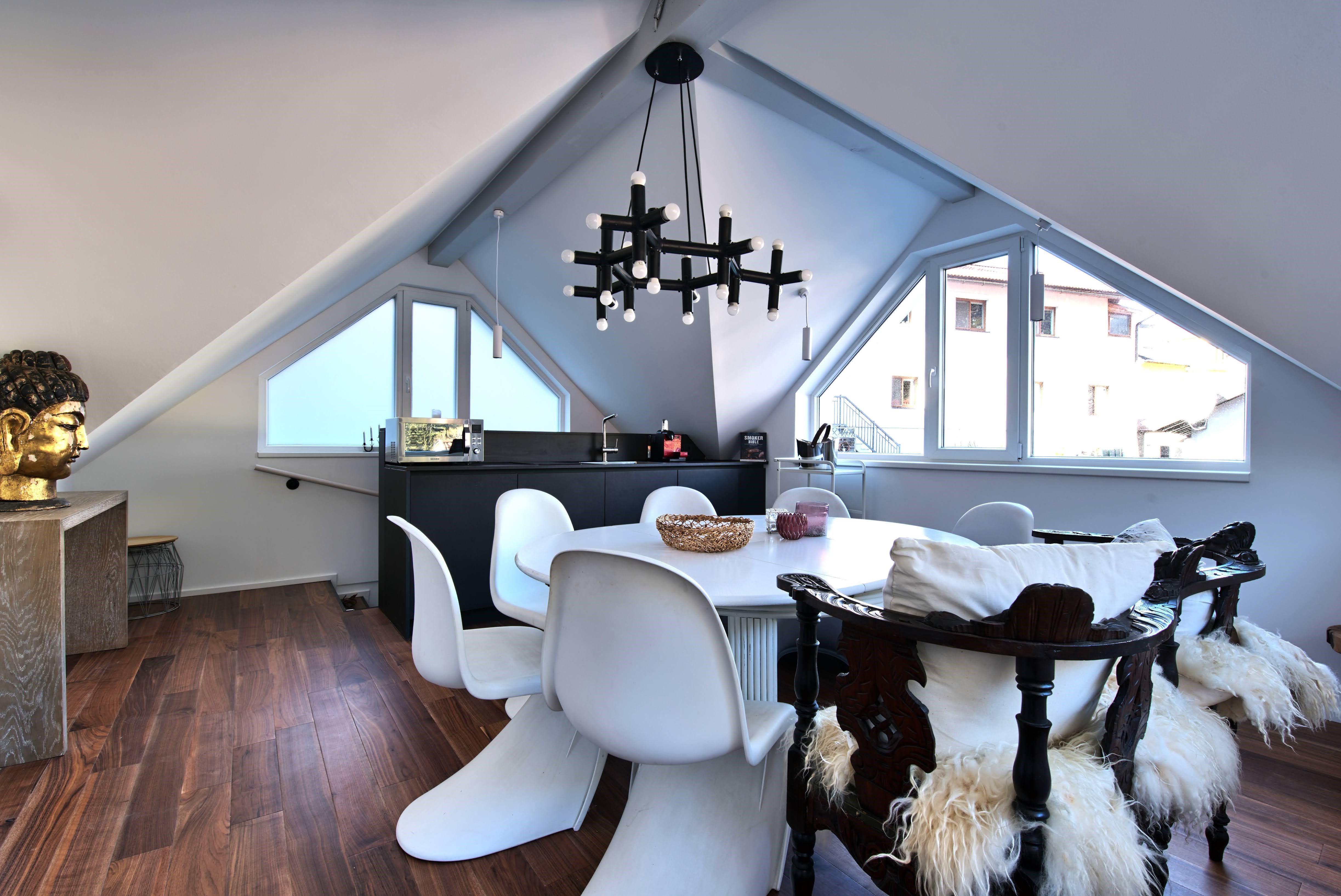 Moderní jídelní kout v podkroví s bílými židlemi, dřevěnou podlahou a velkým oknem.