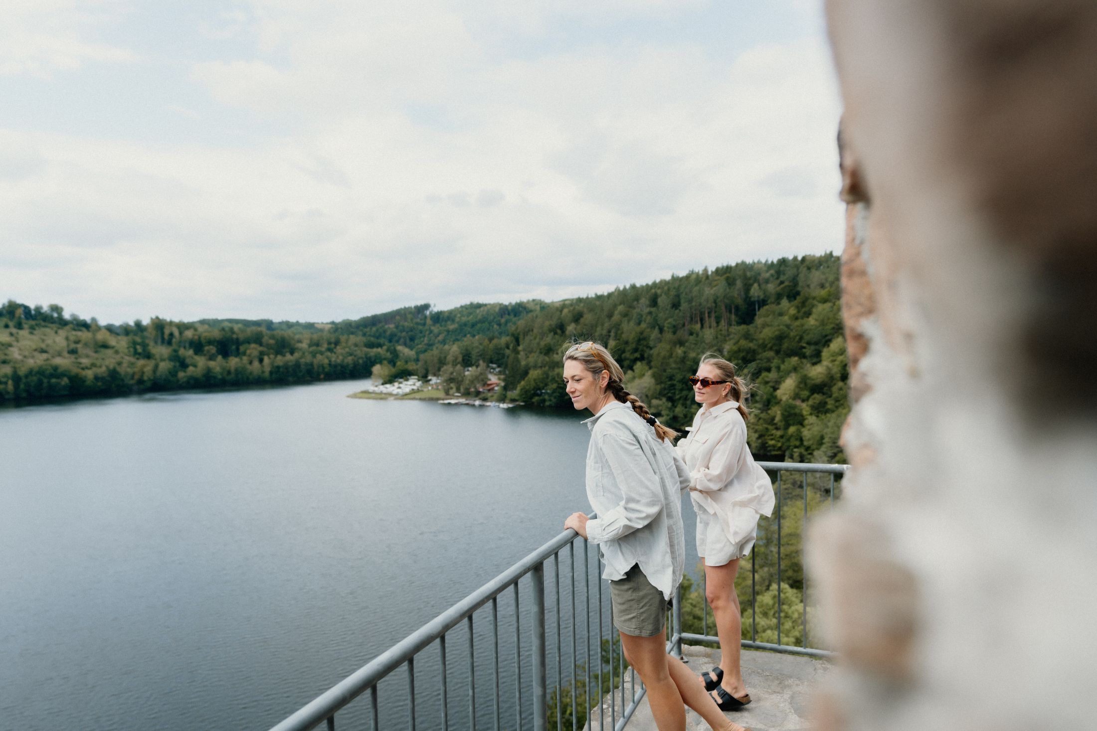 Dvě ženy stojí na balkoně s výhledem na jezero a zalesněné kopce.