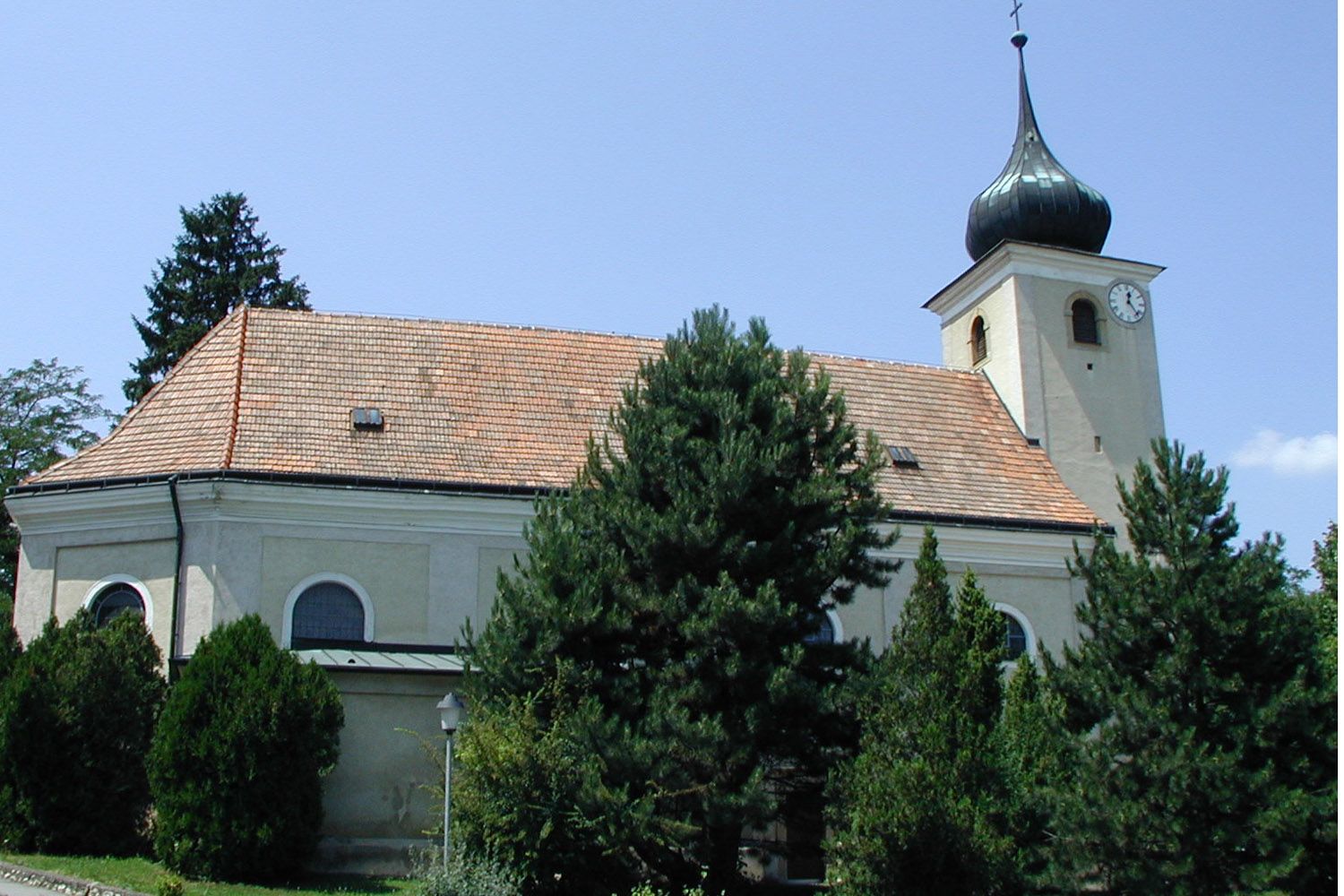 Kostel ve Schleinbachu s věží a hodinami, obklopený stromy.