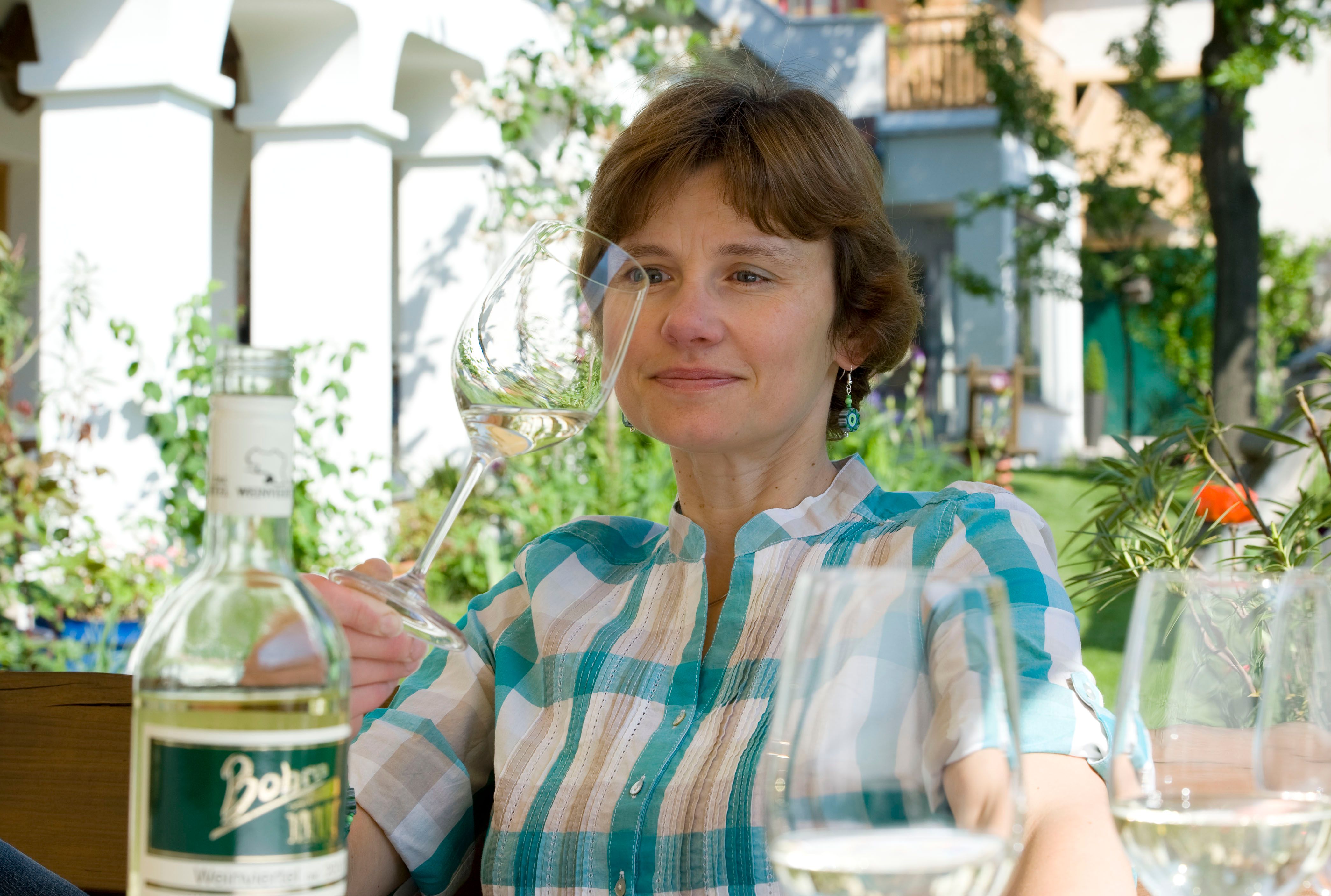 Žena se sklenicí vína na zahradě, v popředí láhev.