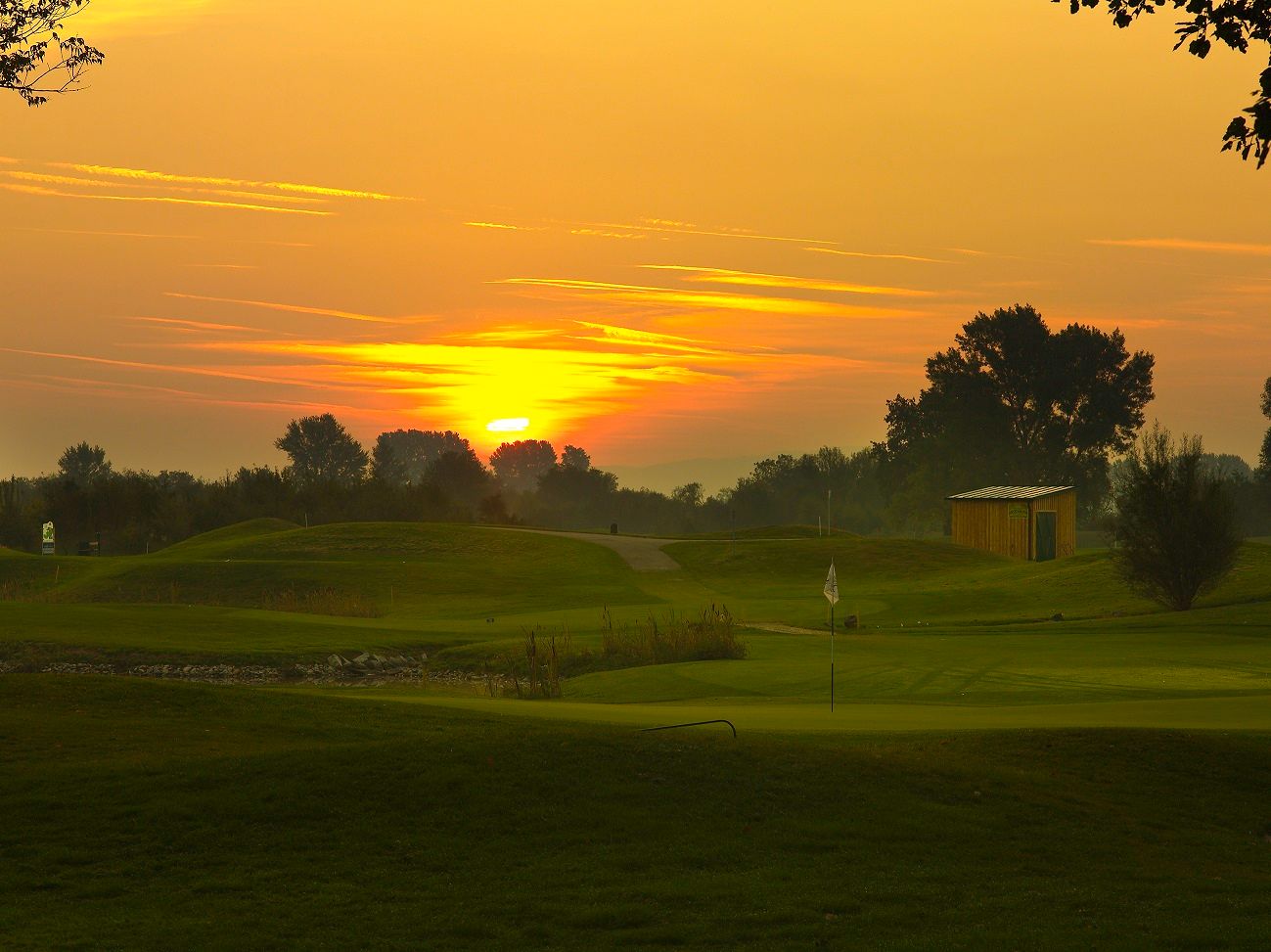 Západ slunce nad golfovým hřištěm s vlajkou a malou budovou.