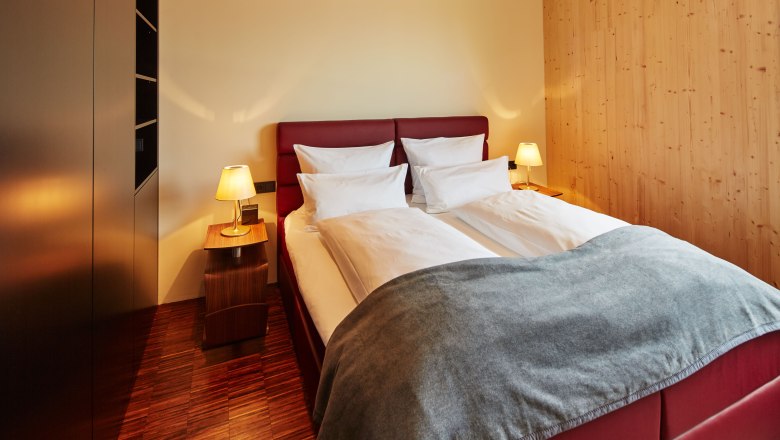 &Uacute;tuln&aacute; ložnice s manželskou postel&iacute;, červen&yacute;m čelem, dvěma nočn&iacute;mi stolky s lampičkami a dřevěn&yacute;mi stěnami.