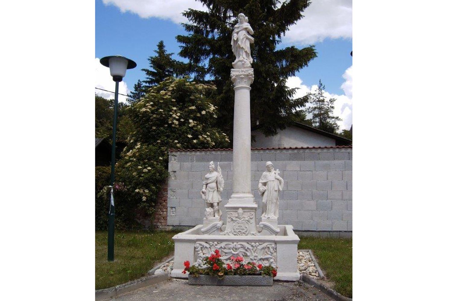 Mariánský sloup se sochami a květinami, obklopený stromy a zdí.