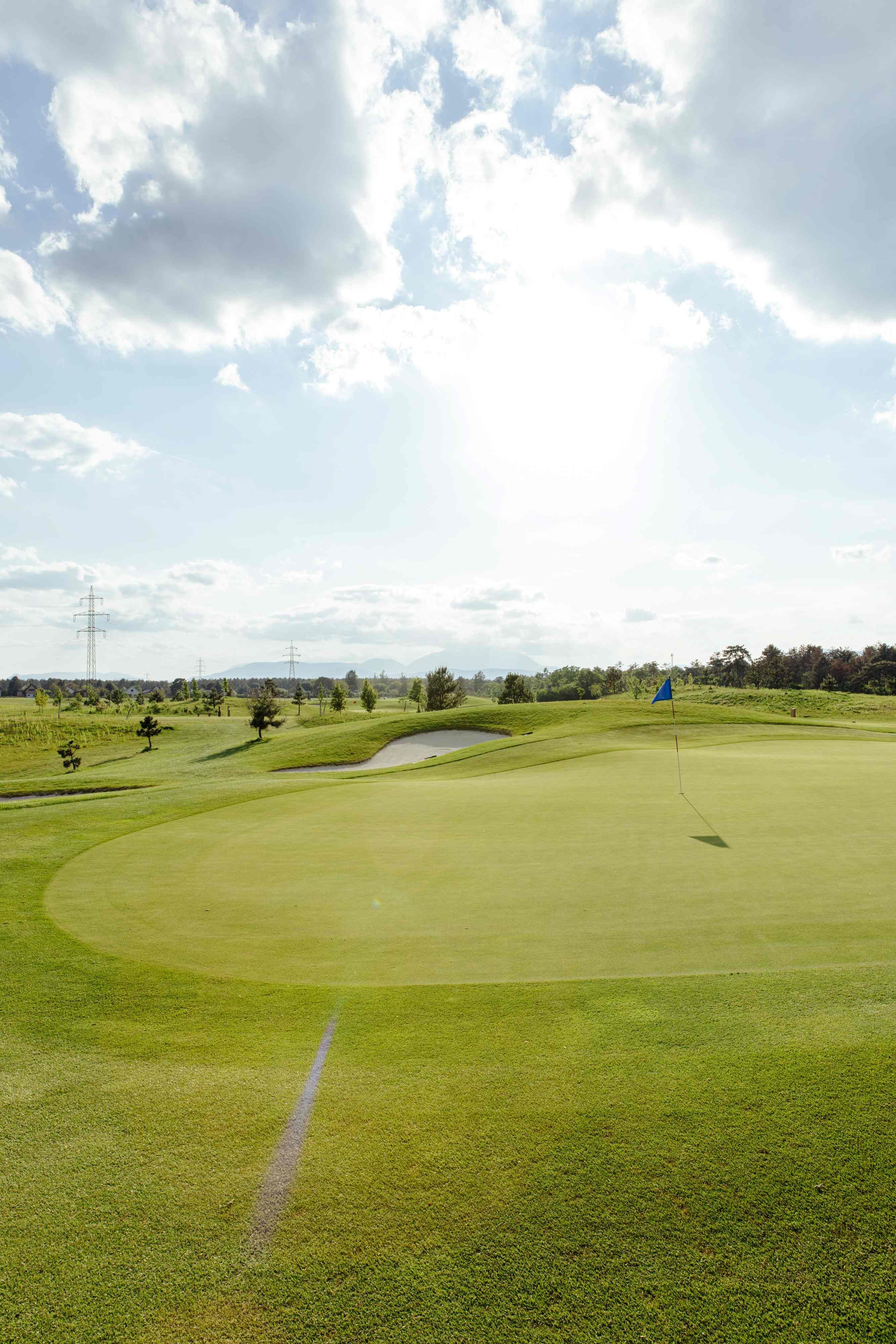 Golfové hřiště s vlajkou na greenu, obklopené stromy a sloupy elektrického vedení pod zamračenou oblohou.