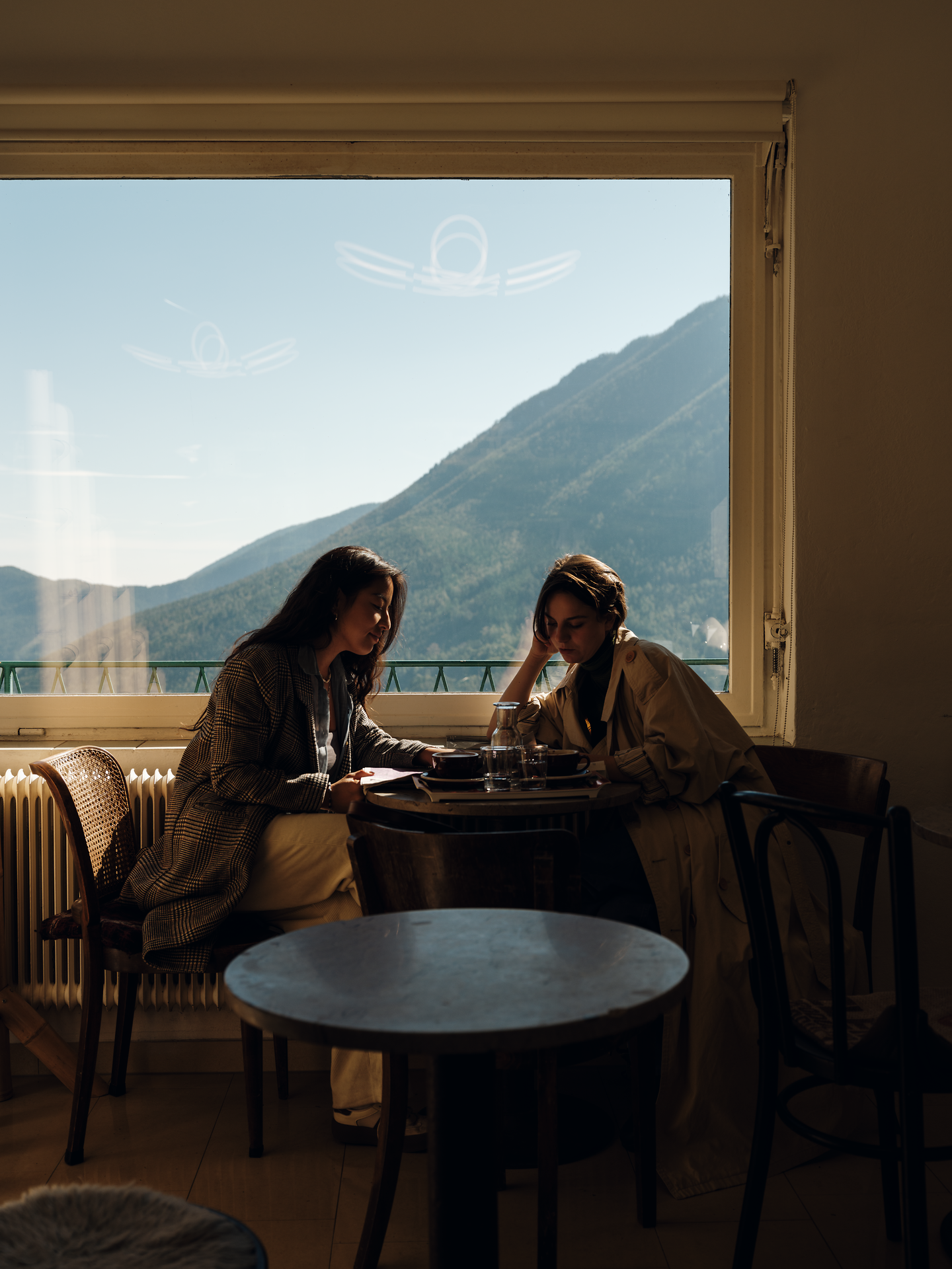 In der gemütlichen Atmosphäre der Kaffeehalle Semmering genießen zwei Freundinnen einen entspannten Moment mit Blick auf die majestätischen Berge. Das sanfte Licht, das durch die großen Fenster strömt, schafft eine einladende Stimmung, während sie sich über ihre Erlebnisse austauschen.