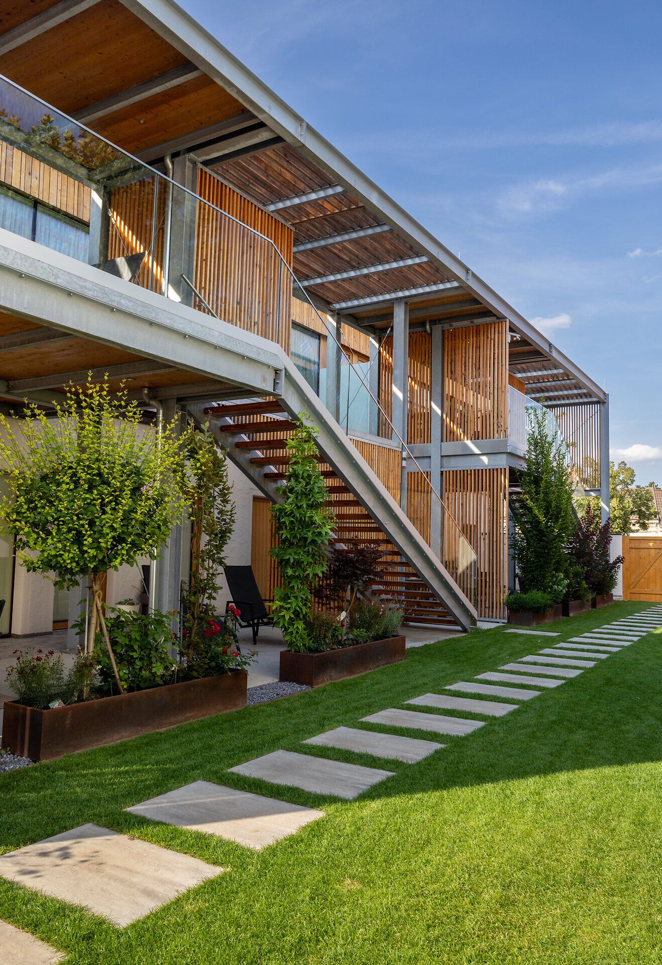 Moderní budova s bílou fasádou a dřevěnými prvky, obehnaná zdí s dřevěnou bránou.